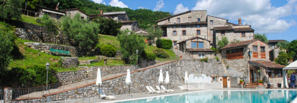 Vista della piscina dell'albergo diffuso Borgo Giusto in Toscana, con le antiche case in pietra della Garfagnana sullo sfondo in una giornata di sole.