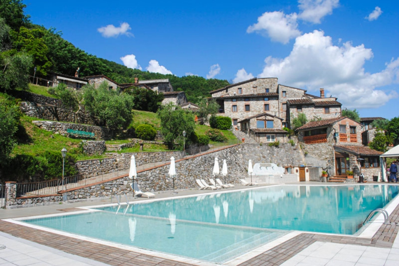 Vista della piscina dell'albergo diffuso Borgo Giusto in Toscana, con le antiche case in pietra della Garfagnana sullo sfondo in una giornata di sole.